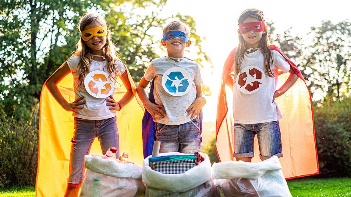 three children standing in front of recycling bins with superhero-inspired masks and capes and the reduce, reuse, recycle symbol on their shirts.